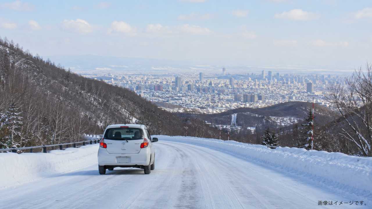 北海道の冬道を走るシンプルな白いコンパクトカー、雪が積もった道路、遠くに札幌の街並み