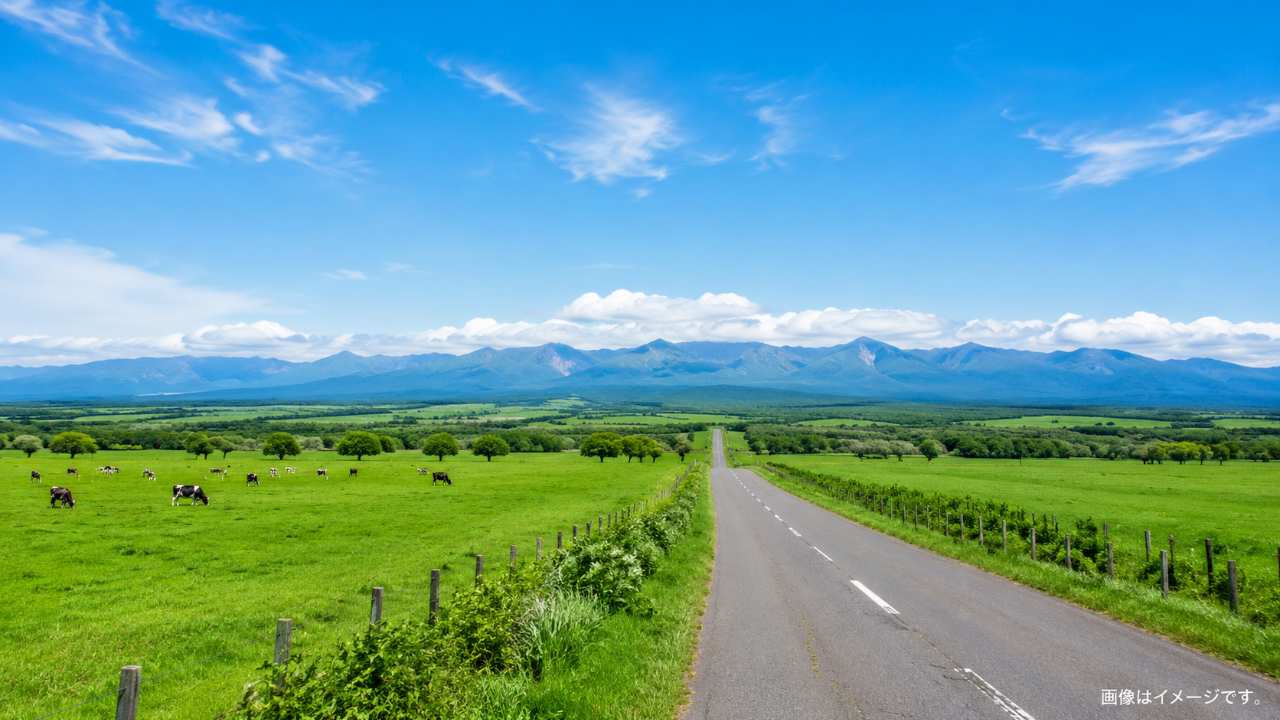 北海道の広大な牧場、遠くの山並み、青空、ドライブロードを写した写真風のブログ画像