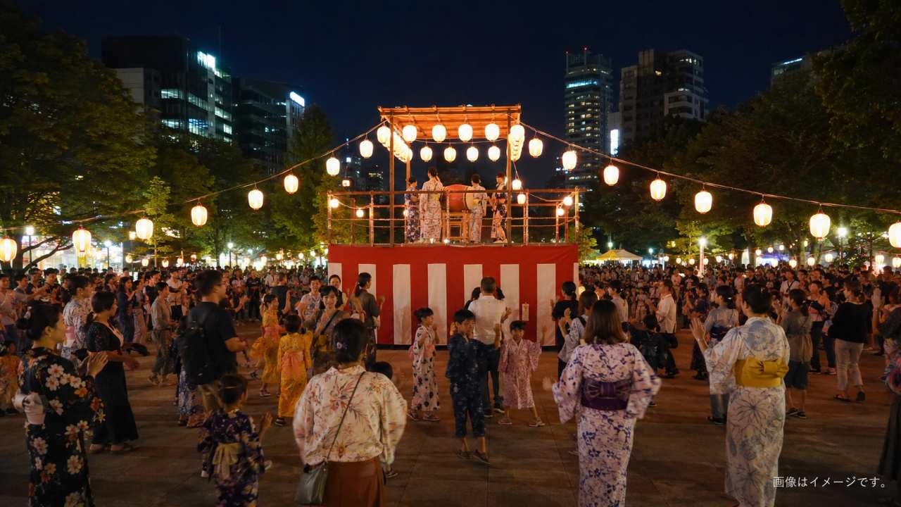札幌の夏の夜、大通公園に設置された盆踊りのやぐらを囲み、浴衣姿の家族や観光客が楽しそうに踊っている